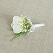 An elegant white orchid boutonniere rests on a softly textured, light beige fabric that looks like finely woven linen, the neutral background allowing every detail of the flowers to stand out. At the heart of the design is a single Phalaenopsis orchid bloom, its broad, velvety petals a pristine, luminous white that curves gently outward, drawing the eye towards the centre where a subtle wash of pale yellow glows in the throat, hinting at freshness and quiet luxury. Surrounding the orchid, a cloud of tiny white baby's breath florets forms a delicate halo, each miniature blossom adding to the sense of lightness and air, like a soft mist around the central bloom. Threaded between the gypsophila are fine strands of feathery green foliage, possibly asparagus fern or another wispy greenery, their fresh, vibrant green tones giving the piece a natural, just-picked feel, as if gathered from a garden near New Barnet's tree-lined streets. The stems are carefully aligned and wrapped in a smooth white satin ribbon, the binding firm yet refined, ending in a small, tidy bow that suggests the steady hands of an experienced florist. Gentle, even lighting falls across the boutonniere, revealing the faint sheen of the orchid petals and the subtle texture of the surrounding flowers without casting harsh shadows, creating a calm and romantic mood. This handcrafted buttonhole feels perfectly suited to a groom or groomsman walking into a New Barnet wedding ceremony, perhaps near Oakhill Park or a local church, where its quiet sophistication and understated purity can complement a tailored suit and capture the promise of a new beginning.