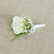 A refined white orchid boutonnière rests on a softly textured, light beige fabric, captured in calm, natural light that emphasises every petal and thread. The focal point is a single, fully opened white orchid bloom, its broad, velvety petals perfectly arranged and gently curved, holding a hint of buttery cream and pale yellow in the throat. This inner glow draws the eye inward and gives the flower a sense of quiet warmth, ideal for a wedding or formal gathering in and around New Barnet. Around the orchid, tiny stars of white baby's breath cluster together, forming a delicate halo that adds volume without overwhelming the central flower. Their small, cloud-like blossoms bring a sense of softness and romance, echoing the fine lace of a bridal gown. Slender strands of vivid green foliage trim the lower edge of the arrangement, emerging like gentle brushstrokes of colour that keep the design fresh and modern, the way many grooms along Station Road or near Victoria Park like their florals to feel. The stems are tightly bound, then wrapped in smooth, pearl-white satin ribbon, creating a sleek handle that would sit comfortably against a tailored suit. A neat satin bow is tied close to the base of the blooms, its loops crisp yet understated, adding a final note of classic elegance. The neutral, woven background fabric, with its subtle weave and linen-like character, frames the boutonnière without distraction, allowing the white tones to glow softly. The image conveys thoughtful craftsmanship and gentle celebration, a timeless accessory ready to be pinned on just before the vows are spoken.