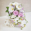 A sophisticated floral centerpiece, ideal for a New Barnet wedding table or elegant hallway console, radiates calm beauty against a softly textured beige background. The arrangement is built around generous stems of white Phalaenopsis orchids, each flower floating outward like a delicate butterfly, its pristine petals gently framing a golden-yellow and orange throat that glows at the heart. Between these orchid sprays, lush lavender-pink roses open in perfect spirals, their petals unfurling in tight, romantic layers that suggest the scent of a spring garden along Lyonsdown Road. Wisps of tiny white baby's breath peek out between the larger blooms, light as lace, softening every transition and filling the spaces with a dreamy, cloudlike feel. Slender stems of light purple lisianthus with slightly darker violet edges add a painterly touch, while cascading sprigs of pastel lavender-pink blossoms-likely stock or sweet peas-trail gracefully down, giving the composition a gentle sense of movement. Fresh green foliage, some leaves slim and tapering, others broader and glossy, creates a verdant frame that makes the pastel petals appear even more luminous. The flowers are gathered into a distinctive white vase formed from interwoven strips that curl into an airy, spherical cage, echoing the contemporary style often seen in stylish New Barnet interiors. At its base, two coordinating woven spheres in pale tones rest on the clean white tabletop, reinforcing the modern sculptural look. Bathed in bright, natural light, this arrangement feels both romantic and current-a piece that would suit a ceremony near New Barnet Station just as well as an intimate anniversary dinner delivered by a trusted local florist.