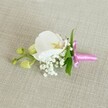 A delicate white orchid boutonniere is laid horizontally on a softly textured, light beige linen-style fabric, its calm, oat-coloured background evoking the gentle neutrals of New Barnet homes and wedding venues. At the heart of the design sits a single, flawless Phalaenopsis orchid bloom, its broad petals a pure, velvety white that curves outward like a small set of porcelain wings. The centre of the orchid glows with soft yellow and the faintest hint of blush pink, adding warmth and quiet depth to the otherwise cool, serene flower. Nestled around the base of the orchid, clusters of tiny white baby's breath create a cloud-like halo, each miniature bloom giving subtle volume and a dreamy, airy texture. A few slim, fresh green leaves peek out behind the flowers, their smooth surfaces and tapered shapes offering a vivid contrast to the white petals and the pale fabric beneath. To one side, several tightly closed, pale lime-green orchid buds extend outward on a slender stem, suggesting new life just about to unfold, much like a wedding morning on a quiet New Barnet side street. The stems of the flowers and foliage are neatly bound together with a bright fuchsia pink satin ribbon, tied into a small, polished bow whose sheen catches the light and adds a celebratory pop of colour. The soft, even lighting reveals the fine weave of the linen, the gentle shadows between petals, and the careful craftsmanship of this boutonniere, ideal for a groom, groomsman, or prom guest wanting refined, understated elegance for a special day in North London.