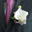 The image captures a refined wedding boutonniere, anchored by a single white orchid that seems to glow against a dark, impeccably pressed jacket. The orchid's petals are wide and velvety, their pure white surface gently warmed by a trace of ivory that softens the overall look. At its centre, a hint of creamy yellow draws focus inward, like a calm heartbeat beneath the flower's serene exterior. Radiating around this focal bloom are cool, silvery-green leaves and delicate lilac blossoms, each stem placed with a florist's practiced hand. Sprigs of gypsophila and pale purple flowers introduce a cloud-like lightness, reminiscent of the blossom-laden trees that brighten New Barnet in early spring. A narrow violet ribbon, tied cleanly around the stems, provides a subtle glimmer of colour; its cool sheen picks up the lilac and pink tones in the tie nestled beneath the lapel. The tie itself, a muted mauve, adds depth and warmth without stealing the spotlight from the orchid. Gentle daylight falls from one side, creating soft shadows in the folds of the suit and the curves of the petals, so that the boutonniere feels almost three-dimensional. The overall impression is one of thoughtful, understated luxury, the kind of floral detail you might choose for a New Barnet wedding celebration close to the High Street or a chic reception near Victoria Park. It is a wearable piece of design that suggests anticipation, commitment, and the subtle excitement of a day when every small element has been selected with care.