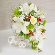 An elegant white and green floral composition is displayed on a sleek white surface, framed by a softly textured beige background that feels as calm and understated as a quiet street in New Barnet at dusk. The centerpiece is formed in a round white wicker basket, its woven structure providing a tactile, handcrafted base that contrasts gently with the smooth petals above. Several large white lilies, some just opening and others fully unfurled, rise from the top, their trumpet-shaped blooms revealing delicate speckling and golden stamens that hint at a subtle, sweet fragrance. Tucked near the upper centre, a creamy pale-yellow rose adds a warm, romantic focal point, its petals layered tightly in a graceful spiral. From the right-hand side, a stream of white Phalaenopsis orchids cascades downward, creating a soft, flowing line that lengthens the arrangement in a style reminiscent of a modern bridal cascade. Around these main blooms, a rich variety of greenery-fine, wispy foliage, slender grasses, and more robust leaves-fills the spaces, giving the arrangement a lush, garden-inspired feel similar to a walk through nearby Oakhill Park. Bright green pom-pom chrysanthemums are nestled among the foliage, their rounded, textured heads adding playful dimension, while delicate clouds of white gypsophila float between the larger flowers, enhancing the sense of lightness. Scattered throughout, glossy ruby-red hypericum berries provide bold, festive accents that stand out vividly against the predominantly white palette. To the left of the basket, two decorative white woven spheres rest on the table, their open, lattice-like design echoing the basket's weave and completing the contemporary look. The overall mood is refined, fresh, and celebratory, perfectly suited for a New Barnet wedding table or an elegant centerpiece delivered to a home near East Barnet Road.
