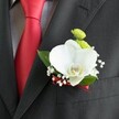 A close-up wedding portrait focuses on a single, meticulously crafted boutonniere pinned to the lapel of a dark charcoal suit jacket, the fine woven texture of the cloth clearly visible beneath the flowers. At the heart of the design sits a large, flawless white Phalaenopsis orchid, its broad petals smooth and velvety, gently cupping a yellow and white lip that glows softly under the light. Tucked just above the orchid, two bright lime-green button chrysanthemum blooms add a cheerful pop of colour, their rounded, cushion-like heads giving the arrangement a playful, modern twist. Around the orchid, delicate sprays of white baby's breath shimmer like tiny clouds, softening the edges and echoing the romance of a wedding ceremony at a local New Barnet church or registry office. Deep green leaves frame the flowers, their glossy surfaces catching the light and grounding the design in natural freshness, as if just gathered from a nearby park in Barnet. A slim, deep red satin ribbon is expertly wound around the stem and secured neatly, perfectly mirroring the rich red satin tie that hangs beside it against a pale grey and white micro-checked shirt. The contrast between the bright red, crisp white, and cool charcoal grey creates a sophisticated, timeless palette, ideal for a groom or groomsman arriving from New Barnet station to a city celebration. Every petal, fold of fabric, and glint of light suggests careful planning and thoughtful artistry, transforming this small floral detail into a quiet statement of love, commitment, and refined style on a day meant to be remembered.