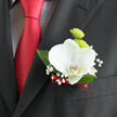 Pinned neatly to the lapel of a dark, well-tailored suit, this orchid boutonnière feels like a gentle signature of style, the kind you might see at a New Barnet registry office wedding or a reception near Victoria Park. At its heart, a single white orchid unfolds with serene grace, its petals broad, silky, and slightly cupped, catching the light like fine porcelain. Subtle warm tones cluster near the centre of the bloom, hinting at soft yellow and cream that deepen its sense of depth. Just behind, two lime-green button chrysanthemums tuck in close, their rounded domes composed of tiny, tightly packed petals that create a playful, textured contrast-bright and lively, like new foliage emerging along the residential streets off Barnet Road in early spring. Fine sprays of baby's breath extend outward, their minuscule white blossoms perched on slender stems, giving the whole arrangement a light, airy border. Deep green leaves frame the flowers, adding body and structure without overwhelming the calm beauty of the orchid. At the base of the design, a deep red ribbon is wound and secured with precision, its rich colour mirroring the sheen of the red tie that runs down the centre of the suit, bringing a hint of passion and formality. The composition feels balanced and intentional, as though every stem has been measured to sit comfortably against the fabric, creating a wearable piece of floral art that accompanies its wearer through vows, photographs, and laughter-filled toasts, carrying with it a sense of New Barnet refinement and heartfelt celebration.