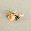 A softly lit close-up of a single peach rose boutonniere rests horizontally on a pale beige, linen-textured cloth, the fine weave of the fabric creating a calm, neutral backdrop reminiscent of an elegant New Barnet wedding venue. The rose is partially open, its petals spiralling gently from a warm apricot centre to creamy, almost shell-like edges, giving the impression of a freshly cut bloom ready for a groom's lapel. Clustered just above the rose, tiny white baby's breath flowers form a delicate halo, their airy blossoms echoing the look of light spring clouds drifting over nearby Oak Hill Park. Slender blades of fresh green foliage, along with two broader, darker green leaves that curl protectively around the base, frame the rose and give structure to the design. The stems are neatly gathered and wrapped in a peach satin ribbon that closely matches the flower's soft tones, the ribbon smoothed carefully by a florist's hands and finished in a discreet, tidy knot. The even illumination across the image highlights the velvety texture of each petal, the crisp whiteness of the gypsophila, and the gentle sheen on the satin wrap. Everything about the composition suggests quiet celebration and thoughtful craftsmanship, as if prepared for a bridegroom heading to a ceremony in one of New Barnet's local churches or registry offices. This floral accessory feels both classic and contemporary, the kind of understated yet refined buttonhole a guest might order for same-day delivery in the area when they want something that whispers romance rather than shouts, capturing the anticipation and tenderness of a special day.