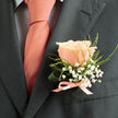 A detailed portrait of formal elegance: a single peachy-pink rose boutonnière rests against the left lapel of a deep charcoal suit, its petals unfolding in finely graduated layers that catch soft daylight and reveal their velvety texture. Surrounding the rose, delicate clusters of baby's breath (gypsophila) create a feathery halo, their tiny white florets floating above slender green sprigs that add a linear, almost calligraphic contrast to the roundness of the bloom. The boutonnière is securely fastened with a pale pink satin ribbon, tied with care into a small, neat bow whose subtle sheen echoes the warm tones of the coordinating tie; beneath this, a pale dress shirt with a faint dark grid pattern peeks through, lending a precise, modern context to the classic floral accent. The photograph emphasises tactile differences - the soft give of petals against the crisp weave of the suit, the whisper of ribbon silk against tailored wool - and suggests the gentle scent of a freshly cut rose, faintly sweet and reassuring. Crafted with obvious attention to proportion and balance, this miniature arrangement feels handcrafted and thoughtful, suited to a groom or celebrant who prefers understated charm. Placing this scene in a local context, one can imagine the finishing touch being pinned on a jacket in New Barnet before a neighbourhood reception by the green or a quiet family gathering, a small botanical promise of celebration and careful craftsmanship.
