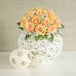 A hand-gathered cluster of peach roses-a wash of honeyed apricot shading to creamy peach-forms a plush, rounded bouquet that reads both contemporary and timeless. Tucked among the roses are delicate sprays of white gypsophila that float like wisps of cloud, softening edges and creating a luminous, ethereal collar; their tiny blossoms scatter light and add a tactile, frothy counterpoint to the velvety rose petals. The arrangement perches within a white woven spherical vessel whose open rattan lattice lends an architectural lightness, allowing glimpses of stems and casting whispering shadows on the surface beneath. Lush green foliage provides depth and a cool contrast, while subtle sunlight-think a gentle morning from New Barnet station toward Brunswick Park-removes any heaviness and highlights the petals' silk-like texture. I can almost smell the faint, sweet perfume of just-cut roses mingled with a green, dewy scent that evokes early walks through local hedgerows. Crafted with care by Flowers New Barnet for occasions that favour understatement-bridal bouquets, intimate gatherings, or a thoughtful gift for a new home-the piece balances artistry and restraint. It reads as a study in quiet celebration: precise yet tender, modern in its vessel but soft in its emotion. Placed on a mantel or a dining table near Barnet High Street, it becomes a gentle focal point that invites lingering glances and soft conversation.
