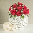 A sophisticated floral display takes centre stage in this scene, perfect for a wedding reception or romantic dinner in New Barnet. A dome-shaped bouquet of deep red roses sits proudly on a white surface, each bloom fully open and velvety, their petals arranged in tight spirals that suggest both freshness and luxury. There appear to be around a dozen to fifteen roses, grouped so closely that they form a rich, textured carpet of crimson. Woven through this sea of red are generous sprays of white baby's breath; their countless tiny blossoms create a delicate mist that lightens the arrangement and lends it an ethereal, almost bridal quality. A few discreet green leaves peek out at the base and between blooms, offering a quiet reminder of the living stems beneath. The bouquet is nestled inside a bulbous white vase with a striking open-weave design. Slim white strands or wicker-like pieces intersect and loop to form a hollow, spherical structure that feels airy and artistic, with glimpses of the stems just visible through the gaps. On the surface to the left rest two decorative woven balls in an off-white or cream shade, one larger and one smaller, their flat, interlaced strips echoing the texture of the vase and creating a harmonious trio of sculptural forms. The background is a soft, neutral wall with a subtle fabric-like texture, keeping all attention on the flowers and their pristine setting. The gentle, even lighting makes the reds appear warm and inviting, much like a carefully prepared table in a New Barnet home ready to welcome guests for an engagement dinner or intimate family celebration.