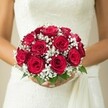 A vision of romance unfolds in this mid-torso photograph of a New Barnet bride, her bridal bouquet a living testament to love and care. The rounded arrangement is dominated by deep red roses, each bloom boasting tightly furled, velvety petals that shimmer with life. Among the roses, wisps of delicate white gypsophila-baby's breath-drift like morning mist, their airy presence offering gentle contrast and a lightness that seems to float above the vibrant green leaves below. Manicured hands gently cradle the bouquet at its base, anchoring the artistry against a backdrop of white lace and soft drape, hinting at the tradition found in local New Barnet weddings. The subtle cream tones of the blurred background recall the peaceful parks and elegant homes of the area, ensuring that all focus lingers on the interplay of color and texture in both dress and bouquet. With every element, from the fresh fragrance of the roses to the peaceful anticipation in the bride's composure, this floral arrangement captures the serene beauty and heartfelt emotion that define a New Barnet wedding celebration.