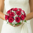 In this intimate wedding scene, a rounded bouquet of red roses and white baby's breath rests gracefully against an ivory bridal gown, as though the bride has paused for a quiet moment outside a New Barnet reception venue. The roses are a deep, classic red, each bloom full and velvety, with petals that spiral tightly at the centre and soften into broader curves at the outer edges. Their surfaces seem almost plush to the touch, catching the light in subtle highlights and shadows that give the bouquet a rich, three-dimensional depth. Threaded between the roses, airy sprays of white gypsophila form delicate clouds, each tiny blossom adding a point of brightness and a lace-like texture, softening the drama of the red while enhancing it at the same time. Slim stems of fresh greenery weave through the design, offering gentle hints of green around the edges, like a quiet nod to the leafy streets that wind between New Barnet and Hadley Wood. The bouquet sits neatly in the bride's hands, tightly bound yet relaxed in appearance, suggesting thoughtful craftsmanship by a local florist who understands the balance between structure and romance. You can almost imagine the faint, sweet fragrance of rose petals drifting on the air as wedding bells echo from a nearby church. This timeless red rose and baby's breath bouquet feels perfectly suited to a heartfelt ceremony in New Barnet, capturing both the excitement of the day and the calm assurance of love that is meant to last.