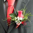 This image shows a handcrafted boutonnière designed with the kind of attentive detail you might expect from a trusted New Barnet flower shop preparing blooms for a wedding at a local church near Station Road. At its centre sits a beautifully formed hot pink rose, the petals gently cupping one another in a graceful spiral, like a tender embrace about to unfold. The colour is rich yet refined, deep enough to convey passion while still feeling appropriate for a sophisticated, formal occasion. Surrounding the rose are airy sprays of baby's breath, or gypsophila, their tiny white blossoms scattered like flecks of snow, creating a soft halo that highlights the rose without competing with it. Fine strands of ornamental grass sweep around the composition in smooth arcs, introducing a sense of flow and rhythm that suggests movement, as if capturing a quiet breeze off nearby Monken Hadley. A single, dark green leaf fans out behind the rose, its glossy surface gently reflecting the ambient light and providing structure and contrast. At the base, a slim red ribbon has been tied into a discreet yet purposeful bow, its sheen echoing the depth of the rose and signalling that this is no ordinary day. The boutonnière rests against a dark suit lapel, the fabric's texture adding a tailored frame that makes the colours appear even more vibrant. The overall impression is one of elegant restraint and heartfelt care, a compact floral accent perfectly suited to a groom, usher, or honour guest wanting a touch of romance that feels both personal and polished.