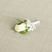 A single white rose boutonniere lies diagonally across a softly textured, light beige fabric, its natural elegance captured as if ready to be pinned onto a lapel for a New Barnet wedding. The rosebud is creamy white with a hint of ivory, its velvety petals still mostly furled, creating a smooth, sculpted dome that speaks of freshness and careful handling. Just at the base, tiny touches of green from the sepals peek through, reminding you this is a real, living flower, likely prepared on the same day for a local ceremony near High Barnet or along the tree-lined streets around East Barnet Road. Surrounding the central rose, delicate clusters of white baby's breath add a cloud-like halo, each miniature bloom forming a tiny star that softens the overall outline and introduces a gentle texture. Slender, deep green leaves and a few feathery, fern-like sprigs cradle the rose, bringing depth and contrast; the greens are arranged to frame the bud rather than overpower it, giving the piece a classic, understated balance that many grooms in New Barnet favour for church or registry office weddings. The stems are neatly bound in a crisp white satin ribbon, wrapped smoothly and finished with a small, precise bow that sits just beneath the flower heads, indicating meticulous florist craftsmanship. The ribbon's sheen subtly catches the soft, diffused light, echoing the gentle highlights on the rose petals. The background fabric, with its fine diagonal weave reminiscent of linen or lightly textured raw silk, provides a neutral but refined stage that suggests a relaxed yet polished celebration. The lighting is even and calming, without harsh shadows, creating a serene mood that suits formal occasions, from intimate ceremonies to larger receptions in local New Barnet venues, and symbolising purity, new beginnings, and quiet confidence in love.