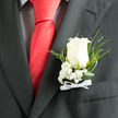 The image focuses on a classic wedding boutonniere pinned to the breast pocket of a charcoal-coloured suit, a familiar sight at New Barnet celebrations from local churches to intimate garden venues off Station Road. At its heart sits a flawless white rose, its petals gently unfurling in a soft spiral that conveys sincerity, devotion, and new beginnings. The bloom's surface appears smooth and velvety, with subtle shadows adding depth and dimension as light brushes across the rose. Surrounding it, airy clusters of white baby's breath cradle the flower, their tiny, star-like blossoms softening the strong line of the lapel. Wisps of fine greenery and delicate fern fronds stretch outward, introducing a natural, slightly wild touch that contrasts beautifully with the sharp tailoring of the suit. The stems are neatly bound and finished with a precise white satin bow, the ribbon's gentle sheen offering a hint of formality and tradition. Together, these elements create a sense of calm elegance, as though the wearer is ready to step into a ceremony at a New Barnet venue, perhaps after photos in nearby parks or quiet residential streets. The arrangement suggests a light, fresh scent-clean, green, and subtly floral-supporting the emotional weight of the moment without shouting for attention. This boutonniere feels thoughtfully composed, the work of an experienced local florist who understands how a single white rose, paired with restrained greenery and careful craftsmanship, can embody the tenderness and quiet joy of weddings, proms, and cherished milestones shared with family and friends.