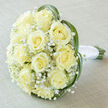Captured from a slightly raised viewpoint, this bridal bouquet centres on a generous cluster of about fifteen to twenty creamy yellow roses, each rose opening into a perfect spiral that forms the lush, domed heart of the arrangement. Wisps of small white Gypsophila are artfully interspersed among the roses, creating a soft, cloud-like texture that offsets the roses' fuller shapes and adds an airy, romantic quality. Around the perimeter, several long, vivid green leaves-reminiscent of bear grass or lily grass-sweep in gentle curves, the arcs tracing an almost heart-like silhouette that cradles the flowers. The lower stems remain visible and are tightly bound with a pristine white satin ribbon; the wrap feels cool and satiny in the hand and is finished with a secure, elegant knot. The bouquet rests upon a pale off-white or light taupe linen surface, its subtle texture providing a neutral, calming backdrop that allows the cream tones to glow. Soft, diffused lighting brings out the faint vanilla blush at the petals' edges and the fresh sheen on the greens, suggesting the quiet intimacy of a spring morning. As the florist at Flowers New Barnet preparing pieces for weddings across Station Road and New Barnet High Street, I picture this bouquet paired with vows exchanged under the green canopies of Oak Hill Park-an emblem of purity, gentle joy, and considered craftsmanship meant for couples seeking something timeless and serene.