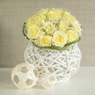 A tastefully arranged wedding table centerpiece is displayed on a glossy white surface, its main feature a generous, dome-shaped bouquet of light yellow roses that appear freshly cut, each bloom gently cupped and fully open. Their soft, buttery petals create a smooth, continuous canopy of colour, while fine sprays of baby's breath are tucked between the roses, their tiny white flowers adding movement and a whisper of texture, like a delicate veil over the arrangement. Around the base, a slim band of bright green foliage wraps neatly, reinforcing the bouquet's rounded silhouette and adding a crisp, botanical edge that suggests careful craftsmanship. This floral composition is cradled in a white woven vase, sculpted from interlocking strands that leave small openings all around, reminiscent of modern wickerwork you might spot in a stylish New Barnet home. The vase's gentle curves and open structure introduce a sense of lightness and contemporary design, complementing the romantic roses without overpowering them. Resting beside the vase are two decorative spheres in soft, natural hues, made from slender strips of rattan- or wood-like material, their open, geometric weave echoing the vase's texture and bringing a subtle, artistic touch to the tabletop. Behind the arrangement, a neutral beige background fades softly out of focus, much like the quiet walls of a New Barnet restaurant set for a wedding or anniversary dinner, allowing the creamy yellows, crisp whites, and fresh greens to stand out clearly. The scene feels serene yet celebratory, perfectly suited to a wedding table arrangement, engagement party, or sophisticated home gathering in and around New Barnet.