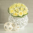 Creamy yellow roses form a compact, domed bouquet in this image, their ruffled centres unfurling like soft sighs and radiating a warm, buttery glow. Interspersed among them, clouds of tiny white gypsophila scatter light and air through the arrangement, the little stars giving the mass a garden-soft texture that contrasts beautifully with the roses' sculpted fullness. The flowers are gathered within a modern white wicker orb, its smooth yet airy weave acting as a sculptural base that lifts the bouquet and introduces a tactile, architectural note. Beside the main vase, minimalist woven spheres offer a playful, contemporary accent-their neutral tone and simple form reinforcing the composition's purity and understated artistry. Smell is almost present: a clean, fresh scent of roses with a whisper of earth, reminiscent of early-summer meadows and freshly cut stems. Curved blades of green frame the bouquet, their looping shapes providing a quiet rhythm and a skilled hand's restraint. Soft daylight from a nearby window highlights the petals' translucence and the wicker's pale grain, creating gentle shadows that enhance depth. As a New Barnet florist I often place arrangements like this in homes near Hadley Green or along Victoria Road where a calm, refined floral statement is wanted-suitable for weddings, a new home gift, or to express heartfelt wishes. Here, the balance of classic blooms and modern vessels shows careful craftsmanship and sensitivity, a piece that feels both soothing and ceremonious without ever being loud.