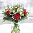 A striking red and white bouquet stands proudly in a clear cylindrical glass vase, its stems neatly trimmed and visible through the fresh water, giving a sense of care and just-arrived freshness, as if it has come straight from a New Barnet flower shop. Deep, velvety red roses are the stars, their petals gently unfurling in perfect spirals that suggest timeless romance and heartfelt passion. At the front, a large crimson carnation with deeply ruffled, layered petals adds dramatic texture and draws the eye, like a richly detailed fabric against the smoothness of the rose petals. Nestled among these bolder blooms are soft white and creamy flowers: elegant white alstroemeria with slim petals marked by delicate speckles, and several tightly closed white lily buds that promise to open in the coming days, extending the life and beauty of the arrangement. Wisps of tiny white gypsophila, or baby's breath, are scattered like a fine mist, lending a cloud-like lightness and generous fullness to the bouquet. Surrounding all of this colour is an abundance of lush greenery. Broad, glossy dark green leaves are artistically curled and wrapped around the base, forming a structured collar that cradles the blossoms. Eucalyptus-like foliage and pointed green leaves in varied tones add depth and movement, echoing the greenery seen in local gardens around New Barnet. The vase sits on a clean white surface against a softly blurred, bright background, perhaps near a window on a quiet Barnet afternoon. Gentle, even light washes across the petals and foliage, highlighting rich reds, crisp whites, and fresh greens, creating an uplifting, celebratory mood. The bouquet feels ideal for anniversaries, birthdays, or Mother's Day, or as a sophisticated gift to brighten a home office overlooking Hadley Green or a cosy front room just off the High Road, expressing joy, appreciation, and warm affection.