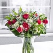 A vivid hand-arranged bouquet of deep crimson red roses and rich red carnations sits perfectly centered in a clear glass vase on a bright white tabletop, captured in soft, natural daylight streaming from a large window behind. Each rose shows off velvety petals, some fully open and romantic, others still in tight, promising buds, while the carnations add ruffled texture and generous volume, echoing the same celebratory red. Between the bold blooms, clusters of white alstroemeria, or Peruvian lilies, appear in different stages of opening; some buds are still green-tipped and closed, others partially unfurl to reveal delicate, trumpet-shaped flowers that lighten the whole composition. Tiny white gypsophila, or baby's breath, is scattered like a fine mist through the arrangement, creating a cloudlike softness that gently diffuses the intensity of the reds. A collar of broad, glossy dark green leaves cradles the flowers at the base, while lighter, more slender greenery threads upward, giving the bouquet graceful movement and depth. Through the transparent vase, neatly cut stems are visible in clear water, a subtle detail that underlines the freshness you might expect from a same-day floral delivery in New Barnet or nearby East Barnet. The white tabletop reflects a faint glow back into the glass, and the background view beyond the window is intentionally blurred and almost monochrome, so that the bouquet's reds, whites, and greens appear to glow with clarity. Overall, the scene feels like a bright, uplifting gift moment-ideal for a birthday, an anniversary, or simply to bring a warm burst of colour into a New Barnet home.
