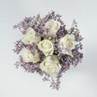 An overhead, high-angle photograph focuses on a compact, perfectly rounded bouquet set against a clean, pale grey surface, allowing every flower to stand out with crystal clarity. At the heart of the design sit six creamy white roses, their petals tightly layered in classic spirals, each bloom at an ideal stage of opening so that the centers are visible but still full of promise. The roses' ivory tones are subtly brushed with the faintest touch of green at the outer sepals, a natural detail that adds freshness and authenticity. Cradling these central blooms is a dense halo of lilac and soft lavender statice or limonium, hundreds of tiny star-like flowers forming a feathery cloud around the roses. The purple tones are muted and gentle rather than bold, creating a cool, serene contrast that makes the white roses appear even more luminous. The texture shifts beautifully from the plush, velvety rose petals to the fine, papery sprigs of statice, suggesting a bouquet that would feel both soft and slightly crisp to the touch. Soft, even overhead lighting casts almost no harsh shadows, enhancing the impression of peace and order, as if the bouquet has just been set down in a calm New Barnet home near Oakhill Park. The image suggests a design created by an attentive local florist, ideal for moments that call for quiet elegance: a sympathy bouquet, a simple bridal posy, or a token of support delivered across New Barnet to bring a sense of comfort and understated refinement.