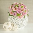 Bathed in gentle, natural light, this artful centerpiece captures the essence of a sophisticated New Barnet celebration-perfect for weddings, milestone anniversaries, or heartfelt gifts. An abundant bouquet of fully bloomed pastel pink and lavender roses sits confidently at the heart of the composition, their plush petals promising softness and subtle fragrance. Delicate pink alstroemeria, with their wisps of yellow and rose-patterned markings, peek out between the roses, embodying the intricate beauty often found in gardens around New Barnet's quiet residential lanes. Pops of lime green button chrysanthemums add a playful, modern accent, while airy baby's breath drifts throughout, lending a feather-light elegance. Dark, glossy green leaves are tastefully interspersed, grounding the arrangement with organic texture. The bouquet is presented in a substantial, round white vase with an intricate open-lattice design-a reflection of New Barnet's flair for combining tradition with contemporary style. To the left, two accent spheres, artistically woven from pale rattan-like strips, complement the vase's craftsmanship and provide a balanced, harmonious tableau. The entire setting is framed by a soft, textured beige background, offering a peaceful counterpoint to the vivid floral hues. This scene is a testament to the artistry and heart of New Barnet's florists-delivering beauty and meaningful moments to every table, from intimate home gatherings to grand celebrations.