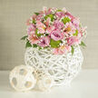 In this image, a rounded pastel bouquet takes center stage, poised on a smooth white table that could easily belong to a light-filled New Barnet apartment near the High Road. The flowers are arranged in a tight, domed shape, every stem carefully placed so that the eye moves slowly over cushions of colour and texture. Soft lavender and blush-pink roses unfurl their velvety petals, some more open than others, creating an interplay of shadows in their spiralled hearts. Nestled among them are delicate alstroemeria in shades of blush and apricot, their long, slender petals swept back to reveal speckled interiors that resemble tiny brushstrokes. Lively lime-green button chrysanthemums stand out with their clean, spherical forms, adding a burst of freshness that keeps the design from feeling too sweet. Wisps of white baby's breath weave through the arrangement, like fine mist over a garden in early morning, softening the transitions between blooms. Dark green leaves appear only in glimpses beneath the flowers, hinting at the natural stems hidden inside the structure. The bouquet rests in a contemporary white woven globe vase, crafted from wicker or rattan, its open lattice criss-crossing to form a sculptural, airy base. Beside it, two lighter, natural-wood decorative spheres echo the same woven pattern, pulling the composition together with quiet harmony. The background is a softly textured, neutral wall, bathed in gentle daylight that enhances every pastel tone. The whole setting feels serene and polished, ideal for a New Barnet wedding collection display or as a romantic centerpiece delivered locally for a special at-home celebration.