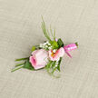 This New Barnet boutonniere is composed like a tiny piece of floral jewellery, centred around a cool pink rose that seems to glow softly from within. Its petals unfurl in fine layers, the outer edges paler and almost sheer, as if lit by soft morning light drifting over The Spires shopping area. Around the rose, blush-toned alstroemeria stretch their trumpet-shaped blooms, each one with delicate yellow centres and thin, painterly streaks of deeper pink, adding subtle contrast without disturbing the harmony. Sprigs of white gypsophila are tucked in like a scatter of stars, filling the spaces between petals and giving the design a light, romantic halo. Slender, fresh-green foliage offers structure, with a few feathery tips and sleek leaves that create a graceful silhouette against the background fabric. The stems are drawn together into a neat cluster and wrapped with a satiny pink ribbon, polished and precise, its tidy finish hinting at the careful hands that crafted it. Resting on woven cloth, the boutonniere feels poised and intentional, as though ready to be pinned on just moments before a ceremony at a local New Barnet venue or a celebration in nearby Hadley Green. You can almost feel the contrast between the rose's velvety touch, the crisp snap of the foliage, and the whisper-light gypsophila. The overall impression is of tenderness and pride-a small floral accent designed to complete a suit with quiet sophistication, perfect for weddings, engagements, or prom nights across New Barnet and the surrounding streets.
