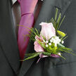 This image shows an exquisitely crafted wedding boutonnière, composed with the keen eye of a florist who knows how to balance tradition and modern style. At its centre rests a pale pink rose, the bloom half-open so that each velvety petal curves outward in a soft spiral, suggesting both innocence and quiet confidence. Around the rose, slender pieces of structured greenery and an elongated blade of foliage give the piece a clean silhouette, echoing the sharp lines of a tailored suit worn to a ceremony in New Barnet or along East Barnet Road. White gypsophila, in tiny clustered blossoms, is woven lightly through the design; the baby's breath forms a cloud of fine texture that softens the strong green shapes and adds a gentle, romantic haze. Tucked near the base is a single green chrysanthemum button, its vivid lime hue and tight, rounded petals introducing a fresh, contemporary energy that keeps the boutonnière from feeling overly traditional. The stems are neatly wrapped in a narrow mauve ribbon, tied so that a slim tail peeks out beneath, giving a subtle, celebratory finish without stealing focus from the flowers. The lighting in the image feels calm and natural, allowing the pastel tones and fresh greens to appear true and lifelike, as if just prepared at a New Barnet flower shop before being delivered for a local wedding. The overall impression is polished yet heartfelt-a small floral accent that quietly reflects care, thought, and the joy of a special day.