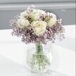 A close-up photograph shows an elegant white and lavender bouquet resting on a smooth, bright white surface, the kind of refined arrangement you might see in a calm front room off East Barnet Road in New Barnet. At the heart of the display are several creamy white roses, their petals perfectly unfurled in soft spirals, with a whisper of pale buttery yellow glowing at the centre of each bloom. The roses look freshly cut, their velvety texture almost touchable, suggesting that they have just arrived from a morning delivery. Around them, clouds of tiny lavender and lilac statice flowers create a gentle haze of colour, their papery, clustered heads forming a delicate, mist-like halo that softens the stronger shapes of the roses. The stems of both roses and statice are gathered neatly in a clear glass vase with a distinctive crackle or ice-effect finish. This textured glass catches the daylight and scatters it in fine reflections across the table, giving the arrangement a subtle sparkle without any harsh glare. Through the water-filled vase, the straight green stems are clearly visible, grounding the airy bouquet with a sense of natural structure and freshness. The background falls away into a softly blurred, pale grey, so that the viewer's eye rests completely on the interplay of white, cream, lavender, and fresh green. The mood is serene and contemplative, like a quiet morning in a New Barnet kitchen, and the design feels ideal for a thoughtful sympathy gesture, a gentle get-well gift, or simply bringing a calming presence to a home office or reading corner. Every detail speaks of measured simplicity, careful craftsmanship, and a love of understated beauty.