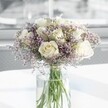 A rounded bouquet of creamy white roses and soft lavender statice stands in the centre of the image, cradled in a tall, clear cylindrical glass vase on a spotless white surface. Each rose is full and perfectly formed, petals gently unfurling from a warm ivory heart to velvety, curved edges, giving the arrangement a feeling of quiet confidence and classic beauty. Around the roses, countless tiny lilac-purple statice blossoms create a hazy cloud of colour, their fine, papery petals adding lightness and texture, like a soft mist drifting around the more structured blooms. The green stems are visible through the glass, neatly aligned and submerged in clear water that catches and reflects the daylight, hinting at the flowers' freshness. Soft natural light spills in from an unseen window, much like a calm morning in a New Barnet living room looking out towards Tudor Park, bathing the bouquet in a gentle glow and casting faint, clean shadows on the table. The background is blurred and airy, suggesting a bright, uncluttered interior where the arrangement becomes the natural focal point. The mood is peaceful and hopeful, suitable for a sympathy gesture, a quiet anniversary at home near East Barnet Road, or simply to bring a touch of serenity to a modern office. Every detail of the composition-the balance of white and lavender, the clear glass, the still water-conveys a sense of thoughtful craftsmanship and understated elegance, as if a local florist has arranged it with care for a meaningful moment.