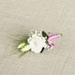Set against a softly textured, light woven background, this boutonniere has the quiet poise of a piece arranged in a calm New Barnet studio just before guests arrive. The central flower is a luminous white lisianthus, fully open, its many ruffled petals folding outwards in gentle curves that suggest both luxury and tenderness. Close beside it, two creamy lisianthus buds lean in, their forms slender and smooth, as if they are just beginning to wake to the day, echoing the feeling of first vows and fresh beginnings often celebrated around New Barnet. Wisps of slender greenery extend from beneath the blooms, offering delicate lines and subtle movement, while sprigs of baby's breath dot the arrangement with tiny white blossoms that resemble a scattering of soft stars. These smaller flowers lighten the composition, creating a sense of airiness and romance without overwhelming the main lisianthus. The stems are carefully aligned and wrapped in a vivid pink satin ribbon, whose shiny surface catches the light with a polished glow. The ribbon is tied into a neat bow that introduces a playful note, like a smile in floral form, contrasting beautifully with the calm white petals and the muted fabric beneath. The overall palette feels fresh, refined, and quietly joyful, ideal for a groom heading to a ceremony near New Barnet station, a prom held in a local hall, or any meaningful event where subtle elegance is treasured. There is a sense of care in every detail, as though a dedicated neighbourhood florist has composed this to sit proudly on a lapel, adding a touch of romance and personality to the wearer's outfit.