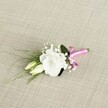 A delicately crafted wedding boutonniere lies at a gentle angle on a light beige, linen-like fabric, its fine weave catching soft, even light like the calm of an early morning in New Barnet. At the heart of the design sits a single white lisianthus bloom, fully open and velvety, its ruffled petals layered in subtle spirals that create a cloudlike, romantic focal point. Just beside it, two slender, pale cream and soft green lisianthus buds rest close to the stem, still closed yet promising to unfurl, echoing the quiet anticipation of a ceremony at a local New Barnet registry office or church. Threaded between the main flower and buds are airy sprigs of white gypsophila, each tiny blossom forming a misty halo that softens the composition and adds a classic, bridal charm. The flowers are supported by fresh greenery: a smooth, darker green leaf cradles the main bloom from behind, while fine, grass-like blades arch gracefully toward the left, introducing gentle movement and a natural, garden-picked feel reminiscent of walks through nearby Oak Hill Park. All the slender stems are carefully gathered and wrapped at the base with a vibrant fuchsia pink satin ribbon, tied into a neat, jaunty bow that brings a modern splash of colour to the timeless white-and-green palette. The contrast between the bright ribbon, the crisp petals, and the neutral textured background makes this petite floral piece ideal for a groom's lapel, a best man, or a stylish prom guest, capturing the quiet elegance and joyful energy that local florists in New Barnet bring to intimate celebrations and same-day event flowers.