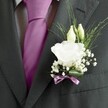 A sharply focused close-up of a groom's charcoal grey suit jacket shows a beautifully crafted white lisianthus buttonhole sitting just above a rich magenta silk tie, the whole look radiating quiet confidence and modern romance. The fabric of the suit has a fine, subtly textured weave, catching the light in soft, matte highlights along the crisply pressed lapel, while a pale grey and white patterned dress shirt peeks out between the jacket and the carefully knotted tie. Pinned to the left lapel, the boutonnière features a single fully open white lisianthus bloom, its many ruffled petals layered like soft tissue, edged in the faintest hint of cream. Two slender, closed ivory buds rise just above the focal flower, suggesting blooms yet to come, and they are framed by airy sprays of white gypsophila, or baby's breath, that create a cloud-like halo around the central blossom. Fine strands of fresh green foliage arc and taper outward, giving the arrangement a light, almost dewy feel against the depth of the charcoal wool. At the base, a broad green leaf curves protectively around the stems, which are neatly wrapped and finished with a narrow mauve ribbon, tied in a small, immaculate bow that quietly echoes the vibrancy of the tie. The composition suggests the calm, anticipatory moments before a wedding ceremony in New Barnet, perhaps on a bright morning near Oakhill Park, where the crisp air, faint floral scent, and immaculate tailoring come together to mark a day of promise, tenderness, and carefully considered style.