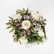 An overhead photograph shows a compact, hand-tied bouquet laid against a pure white background, the clean surface making every petal and leaf stand out clearly. At the heart of the design are creamy white and soft ivory roses, some still cupped in tight buds with a hint of pale yellow at their centres, others fully open so that their spiral of velvety petals can be seen in satisfying detail. Threaded between the roses are stems of alstroemeria, or Peruvian lilies, their slim buds and half-open blooms offering a gentle sweep of mauve, lavender and berry-pink tones. On some petals, fine green streaks and speckles add a painterly touch, like the subtle markings on wildflowers you might spot in Oakhill Park in New Barnet. Tiny gypsophila, or baby's breath, cluster delicately through the bouquet, forming soft white clouds that bring a misty, romantic lightness to the arrangement. Around the flowers, broad, glossy leaves in deep green curve protectively, creating a structured collar that gives the bouquet a sense of polish and calm. The lighting feels even and natural, with no harsh shadows, as if the bouquet were placed near a bright window in a New Barnet home on a fresh spring morning. The overall mood is serene and elegant rather than flashy, making this arrangement perfectly suited to gentle celebrations, quiet anniversaries, or as a thoughtful gesture of support. Every detail in the image speaks of careful craftsmanship and a florist's practiced eye for harmony, colour balance and texture.