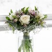 An elegant white rose bouquet stands in a clear glass vase, bathed in soft daylight that feels like it could be streaming through a bright front window in New Barnet. Three creamy white roses are fully open at the centre, their velvety petals gently unfurling to reveal subtle ivory hearts that suggest purity and calm. Around them, smooth, deep green leaves are folded and layered with care, cradling the roses and giving the whole arrangement a rounded, compact shape that feels balanced and serene. Fine sprays of white gypsophila, or baby's breath, weave through the bouquet like tiny clouds, their countless little blooms adding a delicate, almost misty texture. Nestled among the foliage are several alstroemeria buds and blossoms in muted purple and blush-pink tones, some still closed, others just beginning to open, hinting at more colour and life to come. The clear cylindrical vase holds fresh water, and you can see the neat stems gathered together, likely tied just above the waterline with a slim, unobtrusive ribbon that keeps the design tidy. The softly blurred white background enhances the feeling of brightness and peace, allowing every detail of the flowers to stand out without distraction. This arrangement has the quiet emotion of a bouquet you might send to a friend on Lyonsdown Road or near Oak Hill Park, offering comfort, congratulations, or gentle support. The overall impression is of timeless refinement and natural beauty, like a thoughtful piece created by a local florist who understands how to express tenderness through flowers.