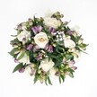 Top-down view of a hand-tied bouquet laid against a pure white background, its circular form glowing with soft cream and deep purple tones. At the heart of the arrangement, several creamy white roses open in layered spirals, their petals shifting gently from warm ivory to pale champagne, like the light over New Barnet just after sunrise. Some roses are fully unfurled, revealing velvety centres, while others remain slightly cupped, suggesting blooms that will open over the next day or two. Threaded between them are clusters of Alstroemeria buds in rich lavender, plum, and magenta, their tapered forms tipped with fresh green at the base, creating a lively contrast against the roses. You can almost trace the delicate streaks that will appear as these Peruvian lilies open, adding subtle pattern and movement. Tiny clouds of white gypsophila float between the larger flowers, their miniature blossoms forming a gossamer veil that lightens the whole design. A ring of glossy dark green foliage frames the bouquet, the leaves peeking out here and there to add depth and a natural, garden-like finish. The lighting is soft and even, without harsh shadows, so every petal edge and vein in the leaves is quietly defined. The overall impression is romantic yet calm, a bouquet that would feel perfectly at home on a wedding table in a New Barnet townhouse or as a thoughtful anniversary gift delivered to a quiet side street off the High Road. It carries a sense of gentle celebration and renewed hope, as if capturing a private, peaceful moment in floral form.