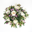 From a bird's-eye view, this sympathy bouquet appears as a perfectly gathered, circular cluster of soft white, gentle purple, and lush green, resting on a spotless white backdrop that makes every colour glow more vividly. At the centre are several creamy white roses, their petals unfolding in generous spirals that transition subtly from pure white at the edges to delicate ivory and a hint of peach nearer the heart, the way warm light pools in a quiet New Barnet living room at dusk. Some blooms are open and welcoming, while others remain tenderly cupped, lending the arrangement a sense of continuity and time passing. Threaded between them, you can see finely poised stems of alstroemeria, many still in bud. These buds show a graceful gradient of tones-from dusky lavender and deeper purple at the very tips down through paler green and blush tones towards the base-like the colour shift you might notice in the borders of gardens along Lyonsdown Road in early spring. A few buds are starting to part at the top, teasing out delicate streaks and striations that promise future blossoms. Throughout the bouquet, small clouds of white baby's breath hover lightly, their tiny blossoms adding an ethereal softness that breaks up the solid forms of the roses. Encasing the flowers, glossy deep green leaves curve around the composition, providing a natural frame and a grounding contrast to the pale blooms. The finished effect is one of composed serenity and heartfelt care, an arrangement that feels right for conveying quiet condolences within the New Barnet community or offering gentle comfort during moments of reflection.