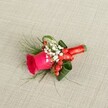 A finely detailed groom's boutonniere rests on softly textured light beige fabric, the weave of the linen or burlap catching the gentle daylight like a subtle backdrop to the flowers. At the heart of the piece sits a deep fuchsia-pink rose, its velvety petals closely furled in the centre and loosening towards the edges, suggesting a bloom picked at perfect freshness from a florist in New Barnet. Around the base of the rose, airy clouds of tiny white baby's breath blossoms create a delicate halo, their miniature petals adding a sense of purity and softness that feels ideal for a wedding morning. Glossy red hypericum berries nestle between the white flowers, their rounded forms and rich scarlet tones offering a playful, festive contrast that hints at winter celebrations or late-autumn nuptials near Hadley Common. Wisps of fine fern-like greenery extend beyond the rose, while broader, darker green leaves are folded and curled to cradle the central flower, giving the design structure and a gentle, sculpted elegance. The stems are neatly bound with a rich red satin ribbon, wrapped smoothly and secured in a precise bow that lies flat for comfort against a lapel. Seen from a slightly elevated angle, every texture is visible: the satin sheen, the smooth berries, the feathery foliage, and the plush rose petals. This handcrafted floral accessory feels suited to a groom, groomsman, or prom date stepping out near New Barnet station, carrying with them a small, romantic statement of joy, care, and celebration on their jacket.