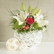A beautifully composed floral display sits on a pristine white surface, styled as the sort of wedding table arrangement often requested for receptions in New Barnet and nearby homes around East Barnet Village. The flowers spill out of a distinctive, round white vase whose open-weave design resembles fine wicker or intertwined branches, giving the impression of lightness and craftsmanship combined. From this textured vessel, tall white lilies emerge with elegance; several blooms are fully open, their broad petals curling slightly at the edges and revealing golden stamens, while a few buds remain closed, their green casings smooth and slender, suggesting that the bouquet will continue to unfold over time. Nestled closely among the lilies is a single standout red rose, its velvety petals forming a neat spiral that lends a focal point of rich, romantic colour, reminiscent of classic bridal flowers. Nearby, bright green pompon-style blossoms add rounded, mossy clusters of texture, bringing a lively contrast to the whites and reds. Wisps of white baby's breath meander delicately throughout, forming a light, airy veil between the stronger blooms. Small clusters of red berries and varied greenery-ranging from feathery, fern-like tendrils to more substantial, glossy green leaves-provide depth, movement, and a sense of natural abundance. To the left of the vase, two decorative balls made from pale, woven strands sit side by side, echoing the vase's open lattice pattern and tying the composition together with a subtle, modern rustic charm. The background is a softly textured, neutral-toned wall, ensuring that every flower, berry, and woven detail stands out clearly. The overall effect is of quiet sophistication and warm celebration, ideal for dressing a wedding table, engagement dinner, or special occasion gathering in New Barnet.