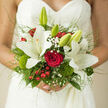 This image shows a luxurious bridal bouquet held against the intricate lace of a wedding dress, the flowers seeming to glow softly as if captured in natural window light somewhere off a quiet New Barnet side street. At the heart of the design rests a single rich red rose, its velvety petals deep and saturated, offering a focal point of passion amidst the surrounding purity. Encircling it, several white lilies open elegantly, their broad, creamy petals spreading into star-like shapes that contrast beautifully with the lace and the bride's skin. A few buds remain gently closed, lending a sense of anticipation and unfolding promise. Between the larger blooms, airy clusters of baby's breath weave in and out, adding a frothy, cloud-like texture. Bright red berries glisten like tiny jewels, their rounded forms and warm tones echoing festive joy, while tufts of mossy green dianthus, mixed with other lush foliage, cushion the flowers in a verdant embrace. Slim strands of ornamental grass arc above and through the bouquet, giving it height and a sense of graceful movement, as if echoing the sway of trees along nearby East Barnet Road. The stems are neatly gathered, likely hand-tied by an experienced florist used to preparing wedding flowers for local churches and registry offices. The entire composition feels balanced and intentional, a harmonious blend of tradition and modern flair, perfectly suited to a New Barnet bride who wants her bouquet to express both romantic devotion and the gentle excitement of stepping into a new chapter of life.