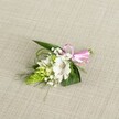 A delicately handcrafted boutonniere is laid out on a softly textured, light beige fabric, similar to natural linen, its neutral tone allowing every petal and leaf to stand out clearly. At the heart of the design sits a single white alstroemeria, or Peruvian lily, its open bloom displaying broad, velvety petals gently ruffled at the edges and finely brushed with reddish-brown speckles near the throat, where the stamens catch the soft light. Just to the left, a tight, lime-green conical spike of unopened buds, reminiscent of Ornithogalum, introduces an intriguing sculptural detail, its scale-like texture contrasting with the smooth lily petals. Airy clusters of white baby's breath weave through the composition like tiny clouds, giving a light, romantic haze around the focal flower. Supporting everything is an artful mix of foliage: strong, dark green, sword-shaped leaves provide structure, while finer grass-like strands and wispy fronds curl and arc, adding movement and depth. At the right, the stems are neatly gathered and secured with a glossy pink satin ribbon tied into a small, precise bow, its cheerful colour bringing a playful note to the refined white and green palette. The lighting is soft and even, as if filtered through a New Barnet morning, revealing the fresh, just-cut quality of the blooms as though they have come straight from a local florist's workbench near Victoria Park. This intimate floral piece feels perfect for a wedding, prom, or anniversary in New Barnet, a small but meaningful accent designed to sit elegantly on a lapel and quietly celebrate a special moment.