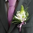 A close-up, high-angle photograph captures the upper torso of a man dressed for a formal celebration in New Barnet, his charcoal grey suit jacket filling most of the frame. The fabric is smooth and sharply tailored, with a clean shoulder line and a neatly pressed lapel that draws the eye to a carefully pinned wedding buttonhole. Beneath the open lapel sits a pale dress shirt patterned with a fine grid, crisp against the skin, and centred by a sleek lavender tie that lends a soft, romantic note to the dark suit. Anchored to the left lapel, the boutonniere becomes the quiet star of the image: a tall, pale green spire of unopened buds rises at the top, its shape slightly tapered, suggesting either young hyacinth or Muscari about to unfold. Clustered beneath it, white alstroemeria blooms open generously, each petal painted with slender maroon streaks and a hint of golden stamens at the heart, giving the flowers a painterly depth. Around them, tiny gypsophila florets drift like a delicate cloud, adding texture and a sense of airiness. Slender, grass-like blades of greenery arc gracefully alongside broader, darker leaves, their rich emerald tones contrasting beautifully with the white petals and the cool purple of the tie. The stems are gathered at the base with a lilac satin ribbon, tied into a polished bow that mirrors the tie's hue and sheen. The lighting suggests soft natural daylight, perhaps just outside a registry office or church near New Barnet station, and the overall mood is one of calm anticipation and understated elegance, the buttonhole quietly marking a milestone moment in the wearer's life.
