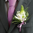 This image shows a carefully composed close-up of formal attire, the sort of detail shot a photographer might take just before a wedding ceremony in New Barnet. The frame is dominated by a dark charcoal grey suit jacket, its lapel pressed to a sharp line and finished with subtle, expert stitching that hints at quality tailoring. Beneath it, a pale shirt with a fine dark check pattern adds quiet interest without distracting from the main focal point: a smooth, solid lavender tie tied in a classic knot, its silky surface reflecting a soft, muted light. Nestled against the left lapel is a beautifully designed boutonniere that appears freshly made by an experienced florist. White alstroemeria blooms, also called Peruvian lilies, open in layered trumpets, their inner petals painted with fine reddish-brown streaks and warm yellow centres that glow gently. From the middle rises a tall, tapering green bud, reminiscent of a tuberose spike, its tightly held petals creating a sleek, architectural form and a crisp lime-green note. Around it dance tiny white stars of baby's breath, or gypsophila, giving the arrangement a light, airy halo. Various greens surround the flowers: narrow, spiky blades, soft, feathery sprigs, and broader, deep green leaves, each providing depth and movement like the foliage in nearby Oakhill Park. At the base, a lilac satin ribbon, perfectly matching the tie's cool purple, is wrapped and tied into a small bow that secures the stems to the lapel. The overall feeling is of cool, poised elegance and quiet anticipation, the kind of floral finishing touch chosen for milestone moments and often ordered for local celebrations through Flowers New Barnet and similar neighbourhood florists.