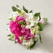 Cradled atop a softly textured beige surface, this hand-tied bouquet glows with the lively character of a New Barnet celebration-ideal for weddings, anniversaries, or heartfelt gestures. Large, luxurious peonies in vivid hot pink immediately draw the eye, their lush, ruffled petals conveying exuberant beauty and a sense of modern romance, perfect for a bride in the New Barnet area. Complementing these showstoppers, clusters of white Alstroemeria add lightness and detailed beauty, each petal adorned with fine maroon streaks and fascinating speckles. Emerging among the main blooms are structural green spikes-perhaps Veronica or early Hyacinth-imbuing the design with texture and playful movement. Smaller, creamy bell-shaped filler blooms nestle into the mix, surrounding the ensemble in a gentle haze, while glossy dark green foliage provides structure and depth. The tight binding of the stems with a brilliant pink satin ribbon denotes both artistry and fresh-from-the-shop perfection, ready for presentation at an event along East Barnet Road or a lush garden gathering near Victoria Recreation Ground. The bouquet's composition, palette, and natural textures together echo both joy and grace, serving as a local symbol of devotion, new beginnings, and the skilled touch of New Barnet's florists.