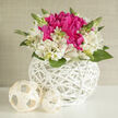 A compact, artfully styled basket of flowers rests against a softly lit background, showcasing the quiet poetry of ivory alstroemeria intertwined with vivid fuchsia blooms. The arrangement is low and rounded, its shape carefully composed so every petal appears to lean into the next, as if the flowers were embracing. At the centre, the deep fuchsia blossoms form lush, velvety clusters, their many layers catching the light and creating pockets of rich shadow. Encircling them, creamy-white alstroemeria open in starry shapes, each petal traced with fine magenta lines that add a painterly detail without overwhelming the design. Slender stems of fresh green, with a few unopened buds, rise gently from the bouquet, suggesting new beginnings and the promise of more colour to come. The flowers are held in a bright white wicker basket, its crisscrossing bands smooth and contemporary yet reminiscent of traditional handwoven work found in local New Barnet markets. Two softly woven cream spheres rest alongside, mirroring the basket's pattern and adding a sculptural note, like minimalist ornaments on a bridal table at a Barnet wedding venue. The lighting is delicate and diffuse, bringing a soft glow to the petals and highlighting the varied greens tucked between them. The overall feeling is of calm celebration and thoughtful generosity, equally suited to a spring engagement party near New Barnet station or a quiet anniversary dinner at home. Every detail suggests care, from the balanced colour palette to the subtle textures that invite a lingering gaze.