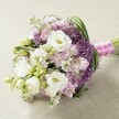 On a softly textured pale beige surface, a lavish, hand-tied bridal bouquet evokes the timeless elegance of New Barnet nuptials. The composition is a harmonious blend of pristine white Lisianthus-frilled petals sheltering creamy yellow hearts-paired with clusters of pastel pink and delicate lavender Stock, their airy textures reminiscent of English gardens in full spring bloom near Friary Park. Punctuating the arrangement are memorable deep violet Allium 'Star of Persia' heads, their starburst clusters adding a playful yet sophisticated architectural edge. Mint-green foliage and bright Lisianthus buds interlace throughout, imparting freshness and dimension much like the natural greenery bordering Barnet's historic lanes. The bouquet's rounded, slightly tapered silhouette is finished with brilliance: a vivid fuchsia satin ribbon, intricately wrapped with luminous white pearls, lending it a romantic, bespoke touch. Soft daylight highlights the bouquet's nuanced colouring and extraordinary textural diversity, drawing out the contrast between the smooth Lisianthus, cloud-like Stock, and striking Allium, all arranged with the expert eye of a New Barnet florist. Evoking feelings of romance, celebration, and hope, this hand-crafted display is perfectly suited for bridal processions, elegant receptions, or inspired gifting across New Barnet, where artistry in flowers celebrates life's most meaningful moments.