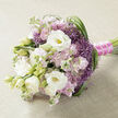 A hand-tied bouquet is presented lengthways on a pale beige, slightly textured fabric, reminiscent of natural linen or fine burlap, forming a calm, neutral backdrop that lets every petal stand out. At the heart of the arrangement, creamy white lisianthus blooms unfold with layered, silky petals, their soft yellow-green centres peeking through like small lights, while several buds remain closed, bringing a sense of life and gentle anticipation. Surrounding them, clusters of soft pink and light mauve stock flowers create an airy, romantic cloud, each tiny ruffled floret adding to the bouquet's fullness and giving the impression of a pastel mist. Threaded between these are striking clusters of purple filler blooms, similar to ornamental allium, their spiky, star-shaped petals forming textured spheres of vivid lavender-purple that provide contrast and a playful edge to the otherwise softly spoken palette. Long, narrow blades of deep green foliage weave in and out of the flowers, some shaped into graceful arcs along the right side, adding structure, rhythm, and a sense of movement, like grasses swaying beside a pathway near New Barnet station. The green tones feel vivid and fresh, echoing the look of just-picked stems from a local garden in Oakleigh Park or Monken Hadley. The stems are expertly aligned and wrapped in a rich fuchsia satin ribbon, smooth and lustrous, which is further adorned with a delicate line of tiny white pearls running along its length, suggesting bridal elegance and timeless style. Crafted with the attention to detail you'd expect from an experienced New Barnet florist, this bouquet speaks of love, celebration, and the quiet joy of holding something beautiful on a special day.