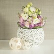 Bathed in gentle daylight reminiscent of a calm afternoon in New Barnet, this enchanting table arrangement captures the artistry and warmth of a neighbourhood florist. An airy sphere-shaped vase, created from open-knit white rattan, cradles a lavish bundle of pastel blooms: creamy white and pale yellow lisianthus unfurl with velvety softness, interlaced with fragrant pink matthiola that adds subtle height and structure. Nestled beneath, deep purple fillers-possibly clusters of aster-bring a pop of energetic colour and delicate texture, while a neat fringe of vibrant green foliage outlines the bouquet, lending a garden-fresh spirit. Flanking the main vase, two light-toned rattan balls with interwoven lattice patterns echo the hand-crafted theme and provide earthy visual interest-a thoughtful nod to the creative flair found throughout New Barnet's shops and studios. Set against an understated beige wall with fine vertical texture, the floral display feels grounded and tranquil, with the clean white tabletop reflecting every soft hue. The overall impression is one of gentle luxury and timeless serenity-evoking the promise of spring, the joy of union, and the calming rhythms of suburban life. Whether enhancing a wedding breakfast in Barnet or brightening a sunlit home, this arrangement from a trusted local florist marries tradition and innovation in every elegant detail.