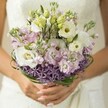 There's a quiet magic in this bridal bouquet-a tapestry of texture and colour uniquely suited to a New Barnet celebration. Held gently in the palms of a bride, the arrangement catches soft afternoon sunlight filtering in, with the elegant white of a wedding gown melting softly into the background. Creamy white Lisianthus blooms, each with gently ruffled petals and hinting at green-yellow centers, sit alongside barely opened buds, conveying both beauty and promise. Clusters of pastel pink and lilac stock flowers cluster in loosely gathered sprays, their multi-petaled forms suggesting carefree joy reminiscent of weekends wandering Hadley Green or window shopping on Barnet High Street. The bottom of the bouquet is accented with a ring of spiky purple Allium, their star-shaped blossoms lending striking contrast and architectural flair-much like the diverse styles of New Barnet's historic homes. Deep, glossy loops of bear grass encircle the base, securing the stems and offering a touch of contemporary design. The bride's neat, softly pink-manicured nails visible at the bouquet's base evoke quiet confidence and anticipation. Every detail, from the gentle transitions of colour to the expertly woven greenery, reflects both the freshness and romantic energy of a spring or summer wedding in New Barnet-a bouquet designed to capture a fleeting, tender moment of joy.