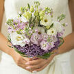 A hand-tied bouquet rests as if poised for a wedding portrait, its palette of lilac, white, and vivid green unfolding like a love story set in New Barnet's quiet back streets. At the heart of the design, white lisianthus blooms open in graceful layers, their petals smooth and slightly curled at the edges, giving a sense of purity and softness. Interwoven between them are stems of stock in pastel lilac and blush, the clustered florets adding fullness and a gentle, nostalgic fragrance that evokes calm walks through nearby Hadley Green on a cool afternoon. Nestled at the base is a ring of lavender allium, their spherical, star-like heads bursting outward in fine points of purple, creating a playful texture that contrasts beautifully with the more romantic, frilled petals above. Encircling the bouquet, glossy green bear grass is carefully threaded and looped, forming delicate spirals that hint at the florist's meticulous craftsmanship, much like the attention to detail in the independent shops lining East Barnet Road. Small, unopened buds in fresh green peek between the open flowers, suggesting growth, anticipation, and the quiet excitement of a ceremony about to begin. The entire arrangement is bathed in soft, natural light, which glances off the smooth petals and highlights subtle tonal shifts from milky white to cool lilac. This bouquet feels both modern and timeless, ideal for brides who want understated elegance for their New Barnet wedding, a design that doesn't shout but instead gently radiates joy, intimacy, and the promise of a shared future.
