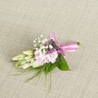 A delicately handcrafted corsage or boutonniere lies centred on a softly textured, oatmeal-coloured linen background, the neutral fabric in gentle, even light that feels like a calm morning in New Barnet. The floral cluster is compact yet full of detail, about the length of a hand, with ruffled soft pink blossoms resembling miniature stocks or garden blooms forming the heart of the design. Their petals shade from a pale blush at the tips into a slightly deeper rose towards the centre, giving the arrangement a romantic, layered look. Threaded amongst these pink flowers are tiny white clouds of baby's breath, each floret no bigger than a pinhead, creating an airy halo that softens the edges. Here and there, discreet sprigs of tiny lavender or deep purple flowers peep through, offering a gentle contrast that keeps the eye moving without overpowering the pastel palette. To one side, several slender, creamy white buds with a hint of pale green-similar to young freesias or delicate lilies-curve outward, still closed and full of promise, as if they are about to open during an evening celebration at a New Barnet venue. A base of smooth, broad emerald leaves cradles the blooms, while fine, wispy fronds of grass-like greenery radiate out, adding a natural, slightly untamed touch. The stems are neatly gathered and bound with a striking fuchsia satin ribbon, tied into a precise bow with short, flowing tails that glimmer softly. The whole accessory feels fresh, cool, and carefully considered, an ideal finishing touch for a wedding, prom, or special family event in and around the quiet streets near New Barnet station.